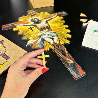 Uma mão feminina com unhas vermelhas segura uma peça de quebra-cabeça de madeira em formato de crucifixo dourado. Ao fundo, o quebra-cabeça maior de Jesus crucificado está parcialmente montado sobre uma mesa preta.