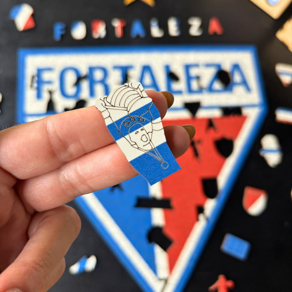 Mão segurando peça de madeira em formato de troféu/torcedor do Fortaleza com o quebra-cabeça ao fundo.