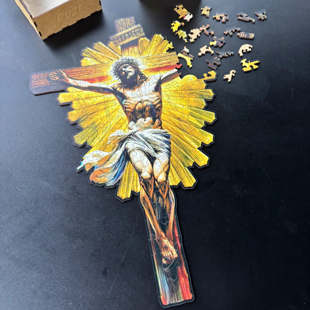 Vista superior de um quebra-cabeça de madeira em formato de cruz com a imagem de Jesus Cristo crucificado. Algumas peças com formatos irregulares e temáticos estão espalhadas ao lado do corpo principal ainda em montagem.